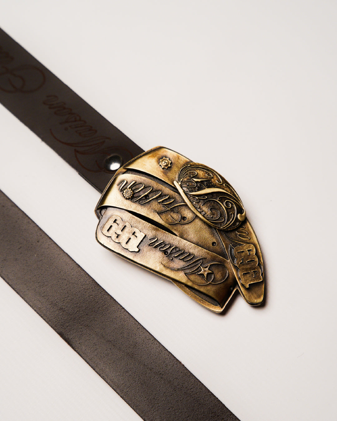 Bronze belt buckle with intricate designs on a brown leather belt against a white background